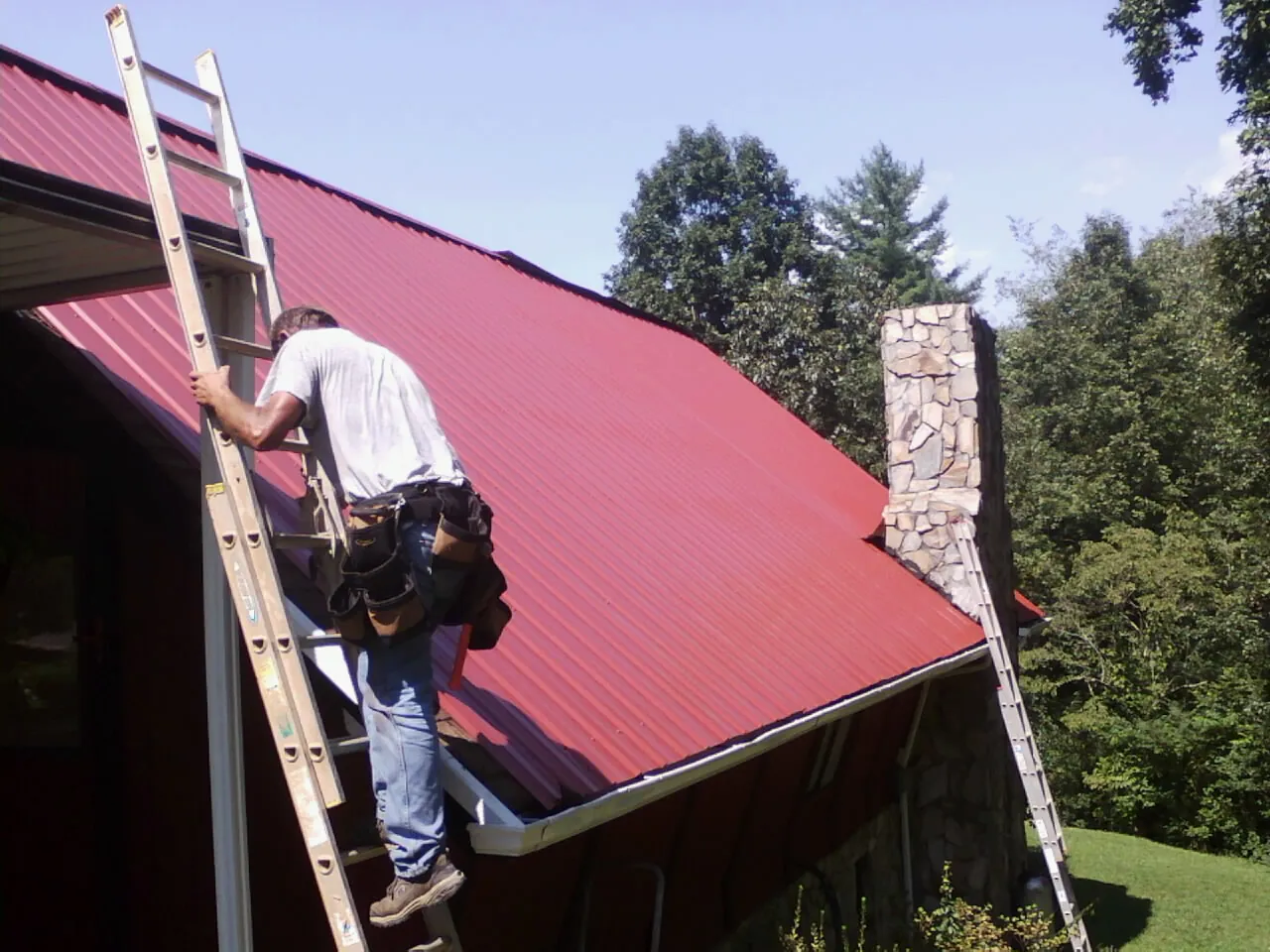 Roofer on a ladder beside a red metal roof and stone chimney — Soffit & Fascia Repair by Roofing Brothers in Breezewood